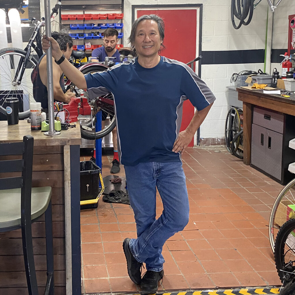 james An Asian man stands in a bike shop with bikes behind him.
